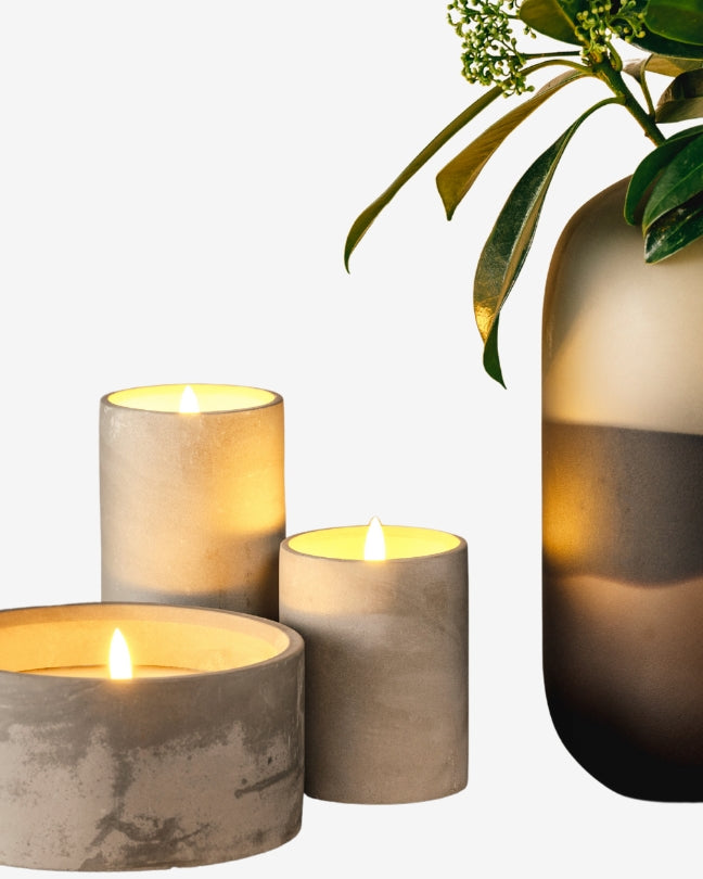 Drei brennende Kerzen im modernen Betonlook stehen neben einer eleganten Glasvase mit grünen Zweigen und verbreiten eine gemütliche Stimmung.