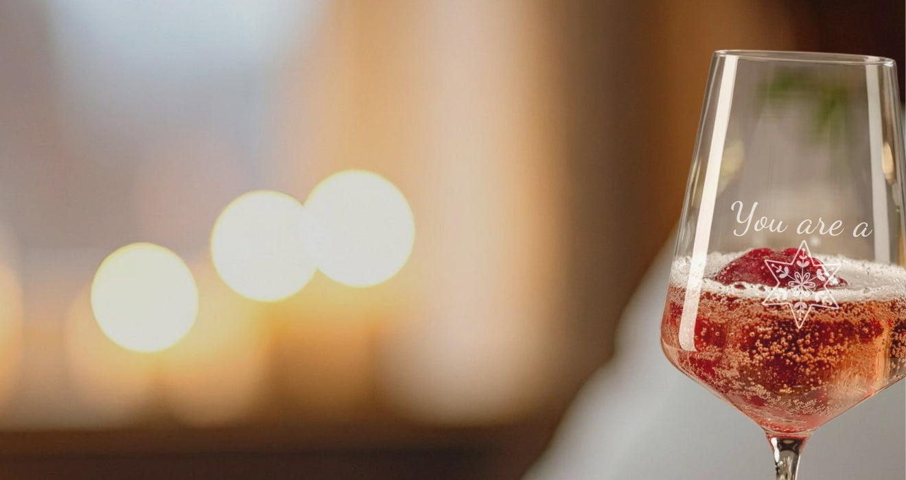 Ein Weinglas mit Gravur „You are a“ und Sternmotiv ist mit prickelndem Rosé und frischen Beeren gefüllt und wird vor unscharf leuchtenden Lichtern in Szene gesetzt.
