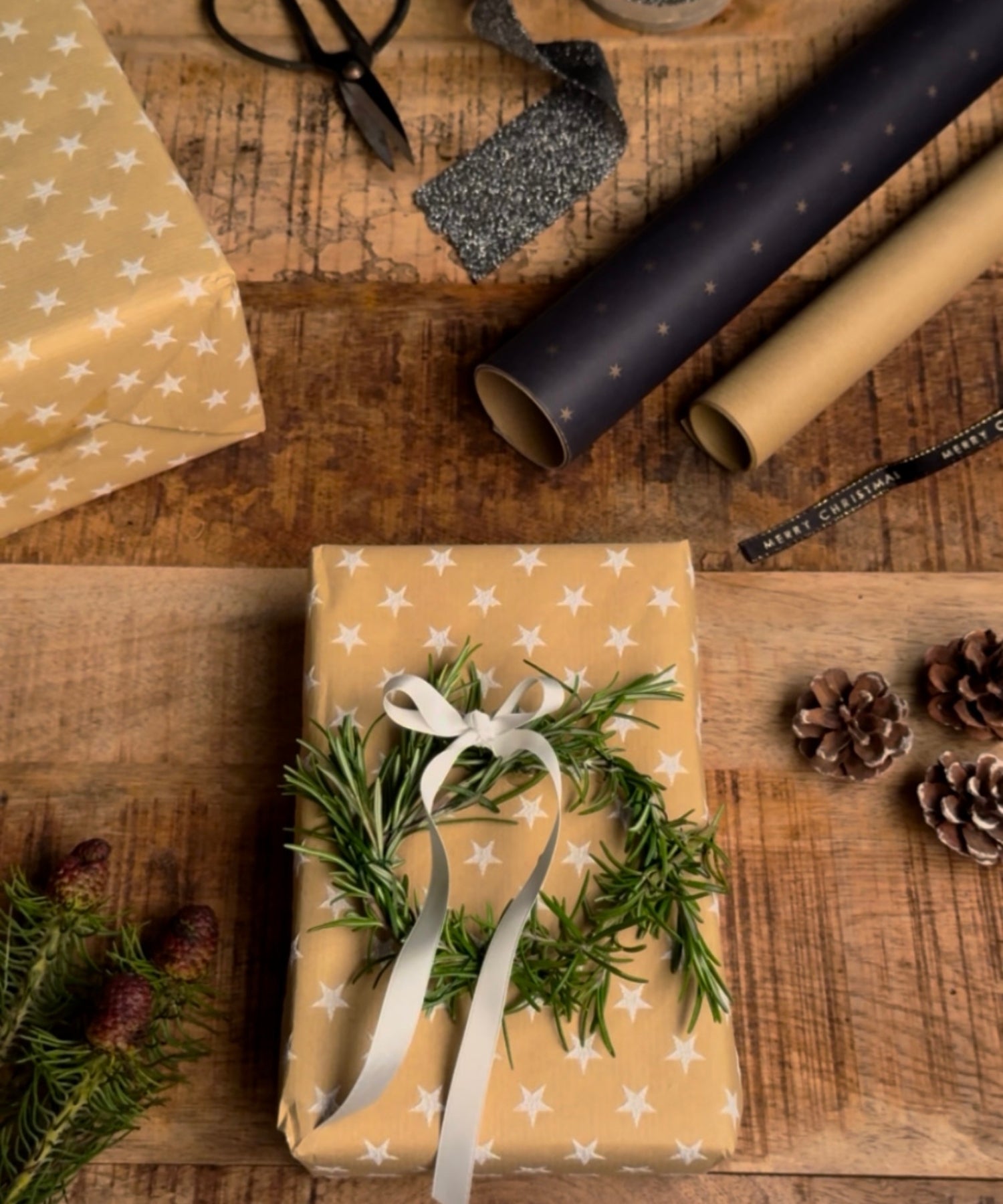 Weihnachtlich verpacktes Geschenk in beige-goldenem Papier mit weißen Sternen, dekoriert mit einem kleinen Rosmarinkranz und weißer Schleife; umgeben von Geschenkpapierrollen, Schere, Band, Tannenzweigen und Tannenzapfen auf einem Holztisch.