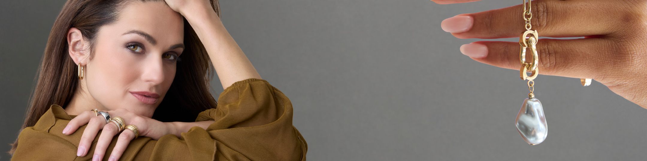 Links im Bild sitzt eine Frau mit goldenen Creolen und auffälligen Ringen, während rechts eine Hand eine goldene Clip-Kette mit großer, schimmernder Barockperle präsentiert – vor grauem Hintergrund perfekt in Szene gesetzt.

