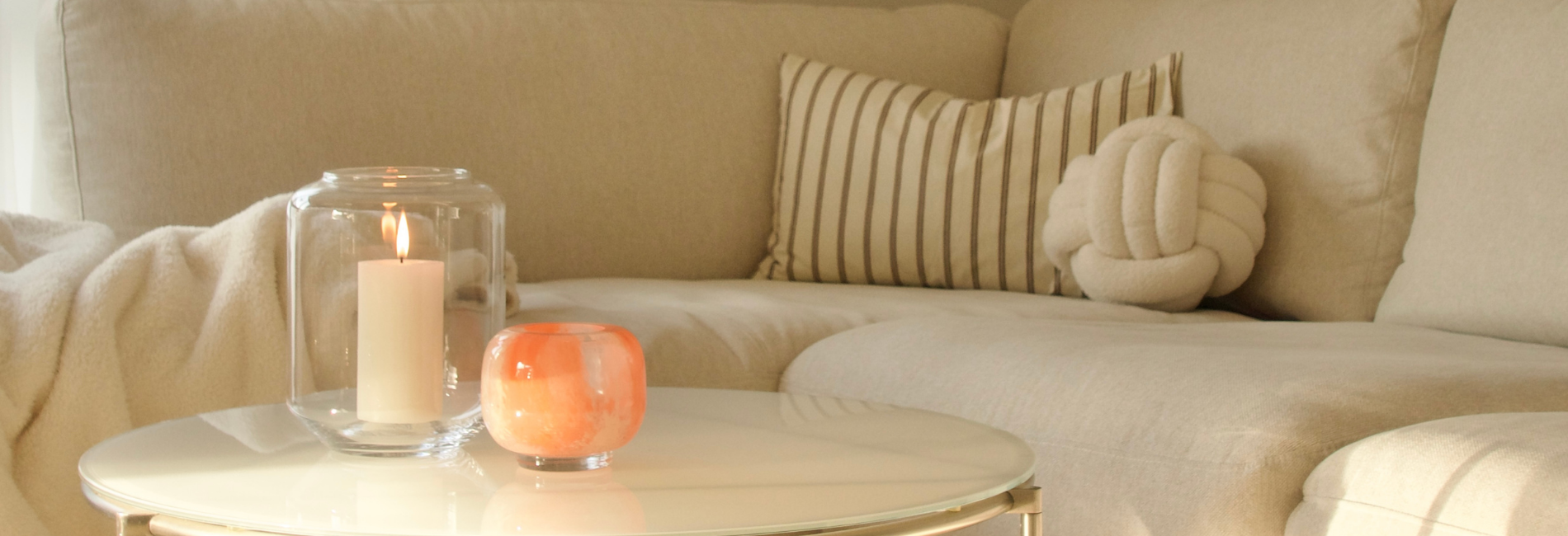 Wohnzimmer mit beigem Sofa, Kissen und Decke; auf einem runden Couchtisch stehen eine Kerze im Glaszylinder und ein orangefarbenes Windlicht.