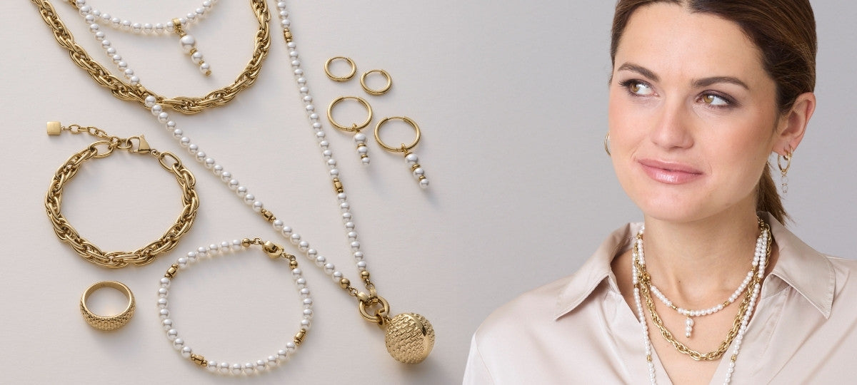 Flach gelegte Schmuckstücke aus Gold und Perlen, darunter Ketten, Armbänder und Ringe. Eine Frau mit beiger Bluse trägt passenden Perlenschmuck – stilvolle Neuheiten-Kollektion.