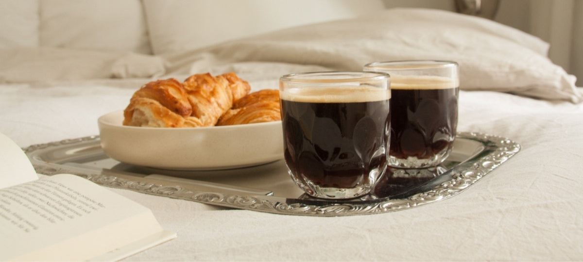 Zwei Gläser schwarzen Kaffee, Croissants in weißer Schale und Buch auf Tablett im Bett mit cremefarbenen Kissen
