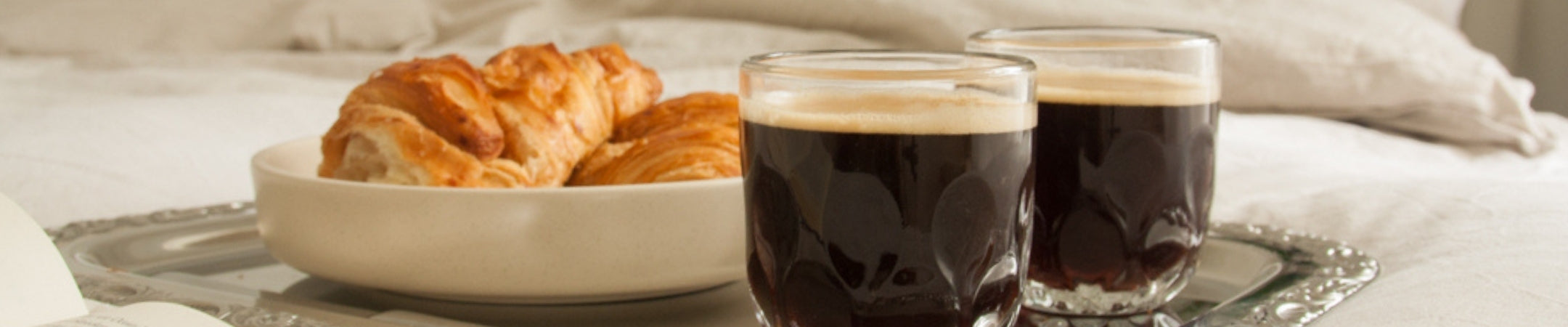 Zwei Gläser schwarzer Kaffee, Croissants in weißer Schale auf hellem Untergrund mit Besteck und Serviette