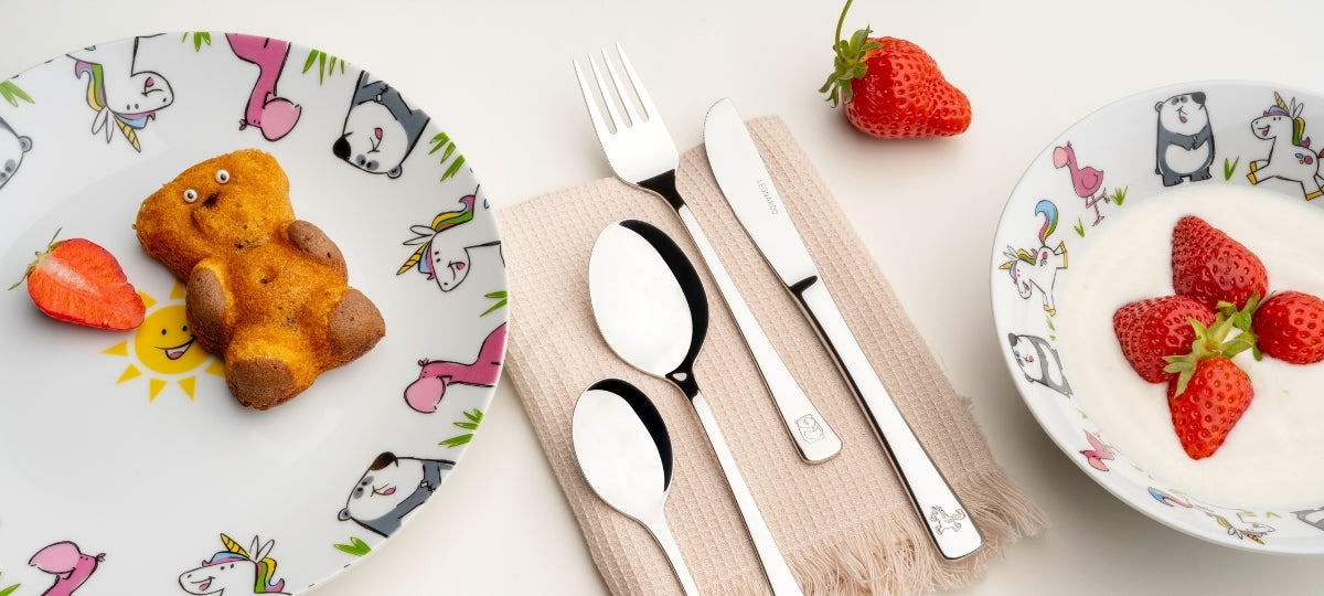 Kinderbesteck und bunte Teller mit Erdbeeren, Teddy-Keks und fröhlichen Tiermotiven auf hellem Untergrund