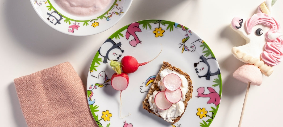 Buntes Kindergeschirr mit Tiermotiven, Radieschen und Lollipops auf weißem Hintergrund, rosa Serviette und Gebäck