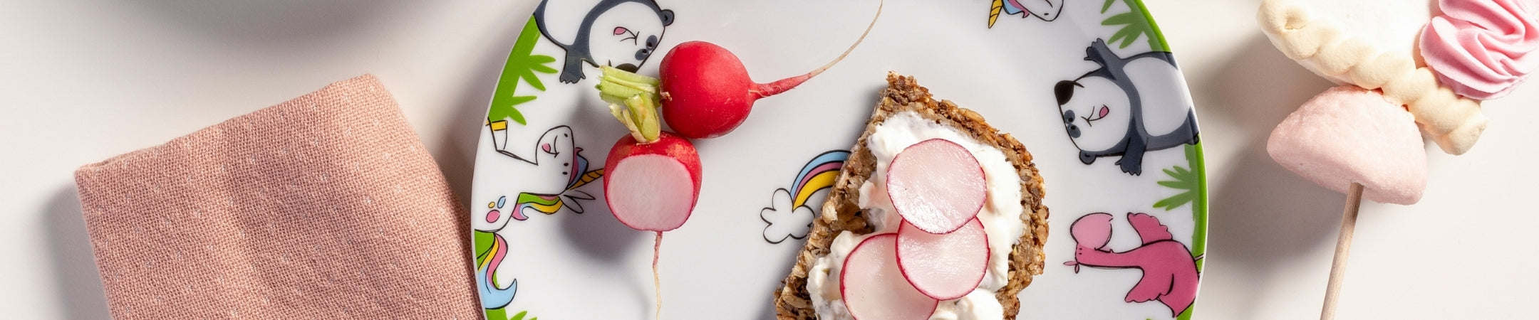 Kindergeschirr mit buntem Muster, Radieschen auf Toast, rosa Handtuch, Marshmallows und Lollipops auf weißem Hintergrund
