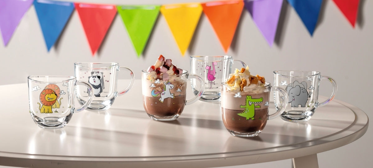 Bunte Kindergläser mit Tiermotiven auf silbernem Tisch, gefüllt mit Kakao und Desserts, bunte Wimpelkette im Hintergrund