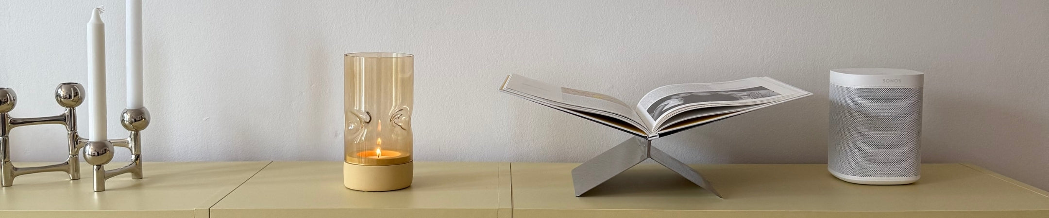 Kerzenständer, goldene Kerze im Glas, offenes Buch und weiße Dose auf hellem Holztisch