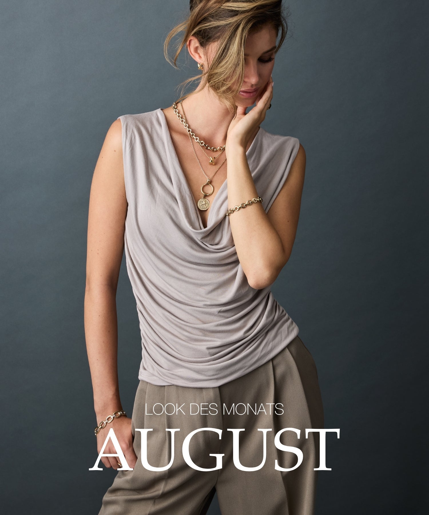 Look des Monats August: Frau mit beigem Wickeltop und Leinenhose trägt goldene Schmuckketten und Armbänder vor grauem Hintergrund