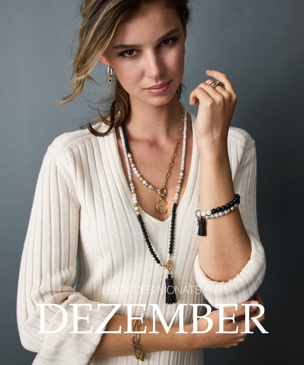 Lookdes Monats Dezember: Frau mit cremefarbenem Strickoberteil, mehrlagigen Perlenketten und Schmuckarmband vor grauem Hintergrund