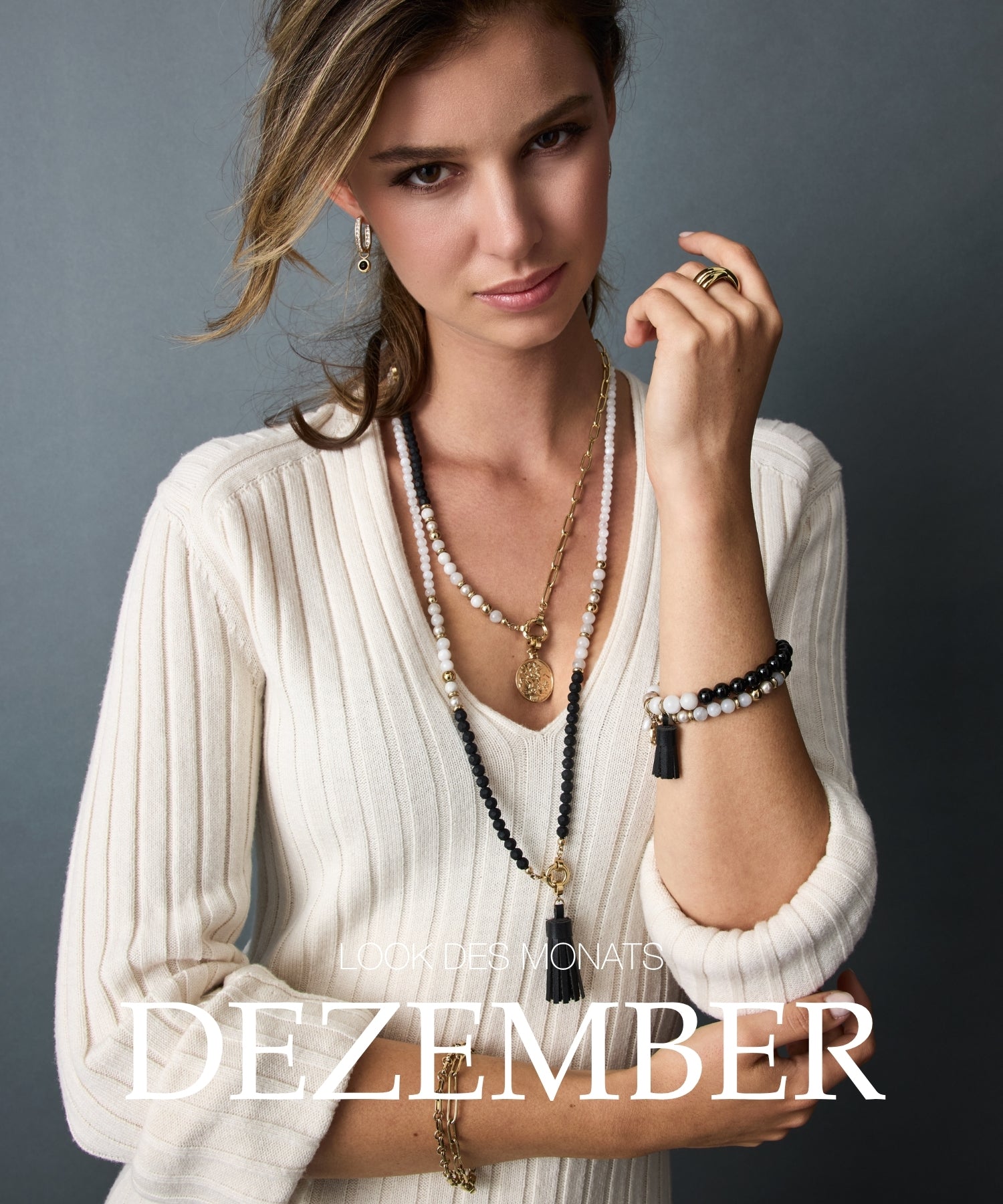 Lookdes Monats Dezember: Frau mit cremefarbenem Strickoberteil, mehrlagigen Perlenketten und Schmuckarmband vor grauem Hintergrund