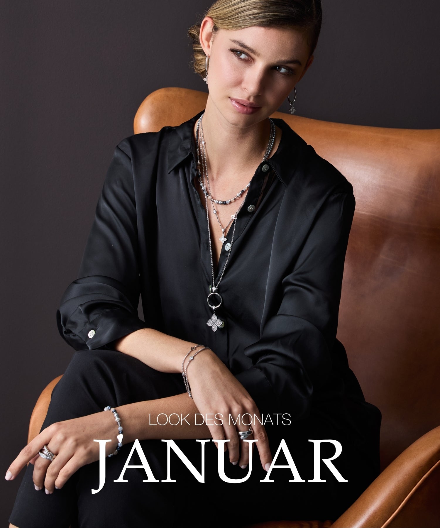 Look des Monats Januar: Frau mit blondem Haar in schwarzem Hemd mit Schmuck und Accessoires auf braunem Ledersessel