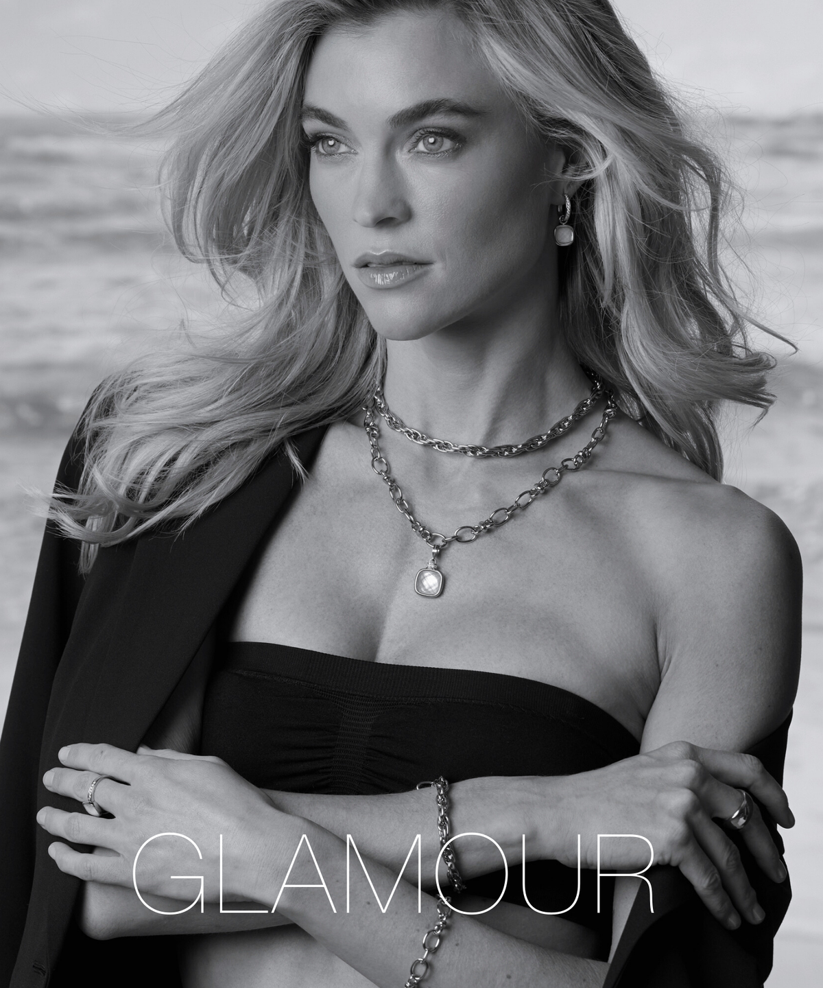 Eine Frau mit offenen blonden Haaren blickt seitlich in die Ferne und trägt eine elegante Kette und ein Armband. Ihr Outfit und der Schmuck wirken edel und stilvoll vor maritimem Hintergrund.