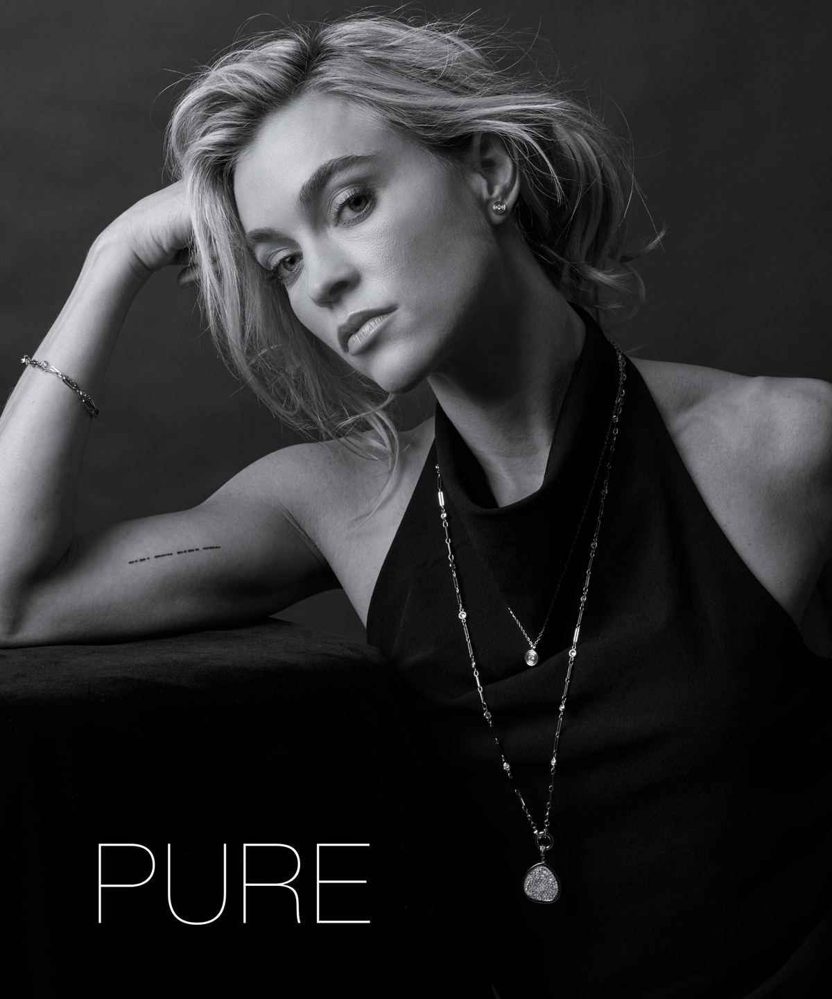 Eine Frau lehnt mit dem Kopf auf dem Arm und trägt eine lange Halskette mit Anhänger. Ihr reduzierter Look und der schlichte Schmuck wirken modern und zurückhaltend.