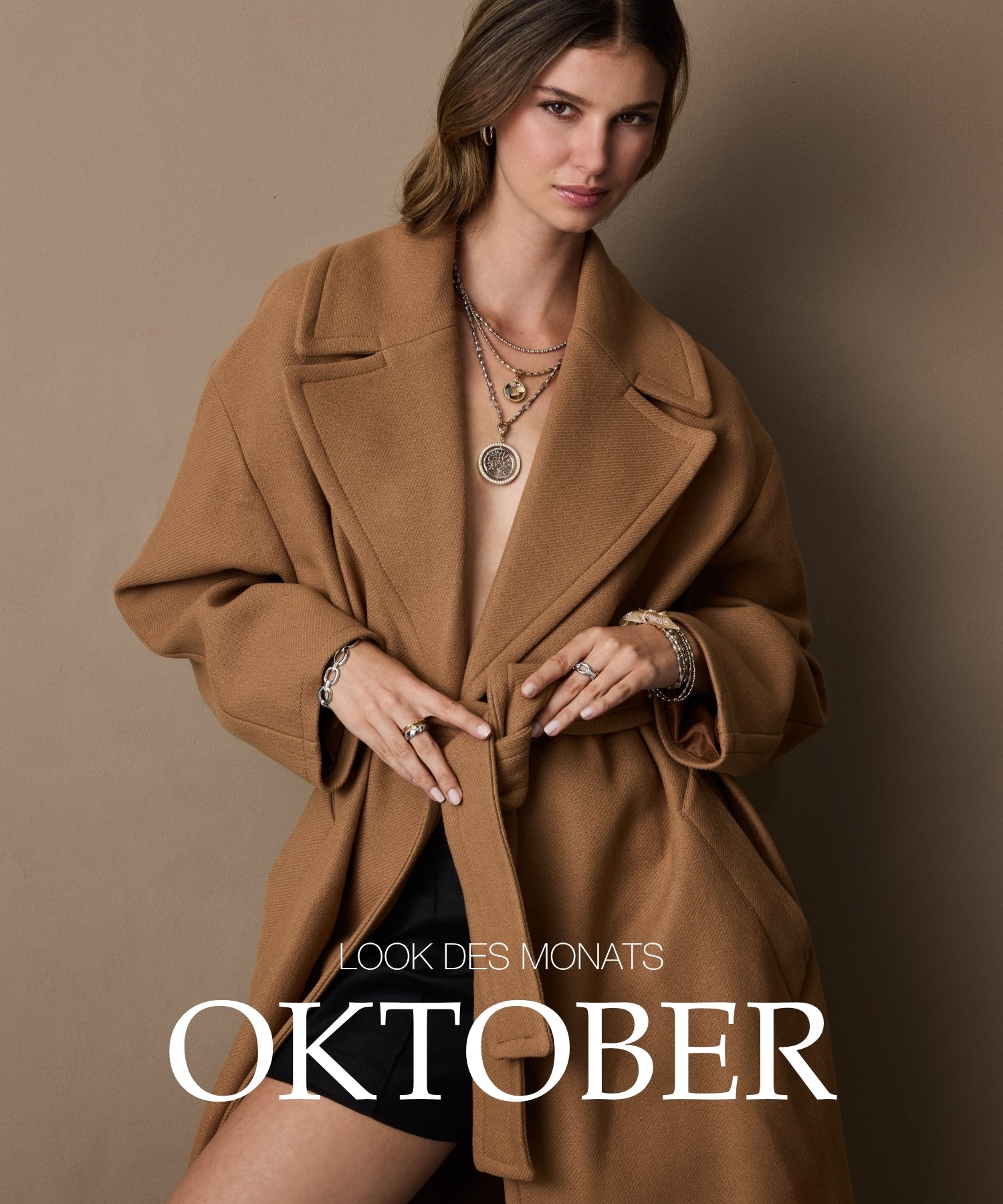 Look des Monats Oktober: Frau trägt camelfarben Wollmantel mit Schmuck und Ringen vor beigem Hintergrund