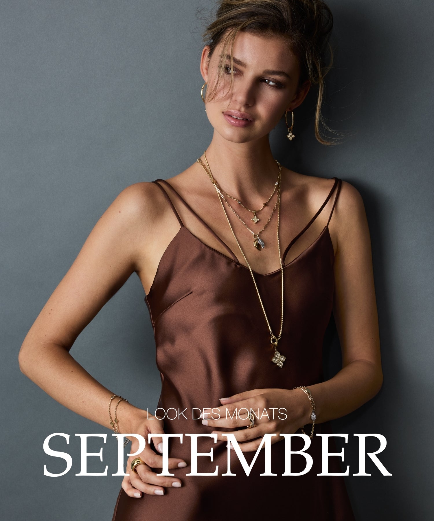 Look des Monats September: Frau mit braunem Seidenkleid und goldenem Schmuck vor grauem Hintergrund