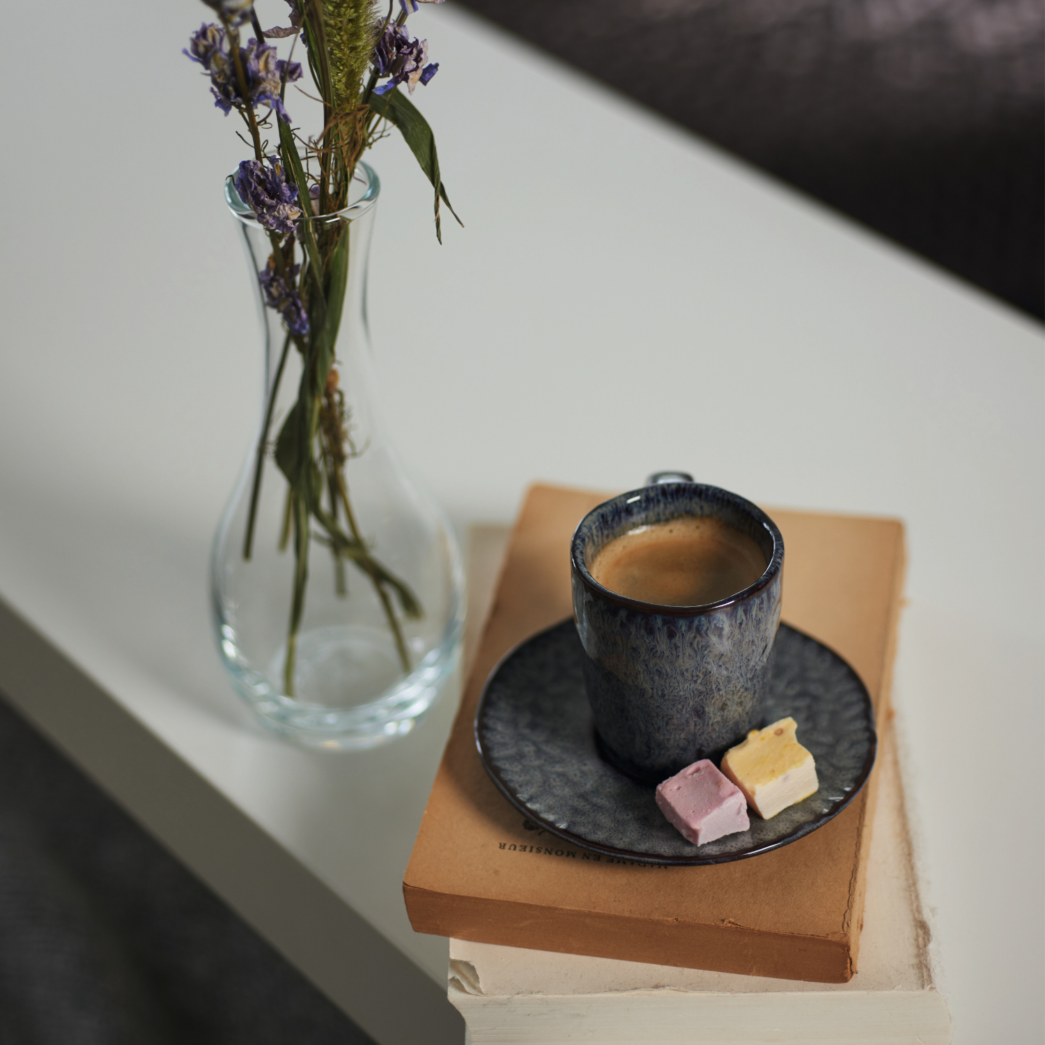 Eine elegante Espressotasse mit Unterteller steht auf zwei Büchern, begleitet von einer klaren Glasvase mit getrockneten Blüten.