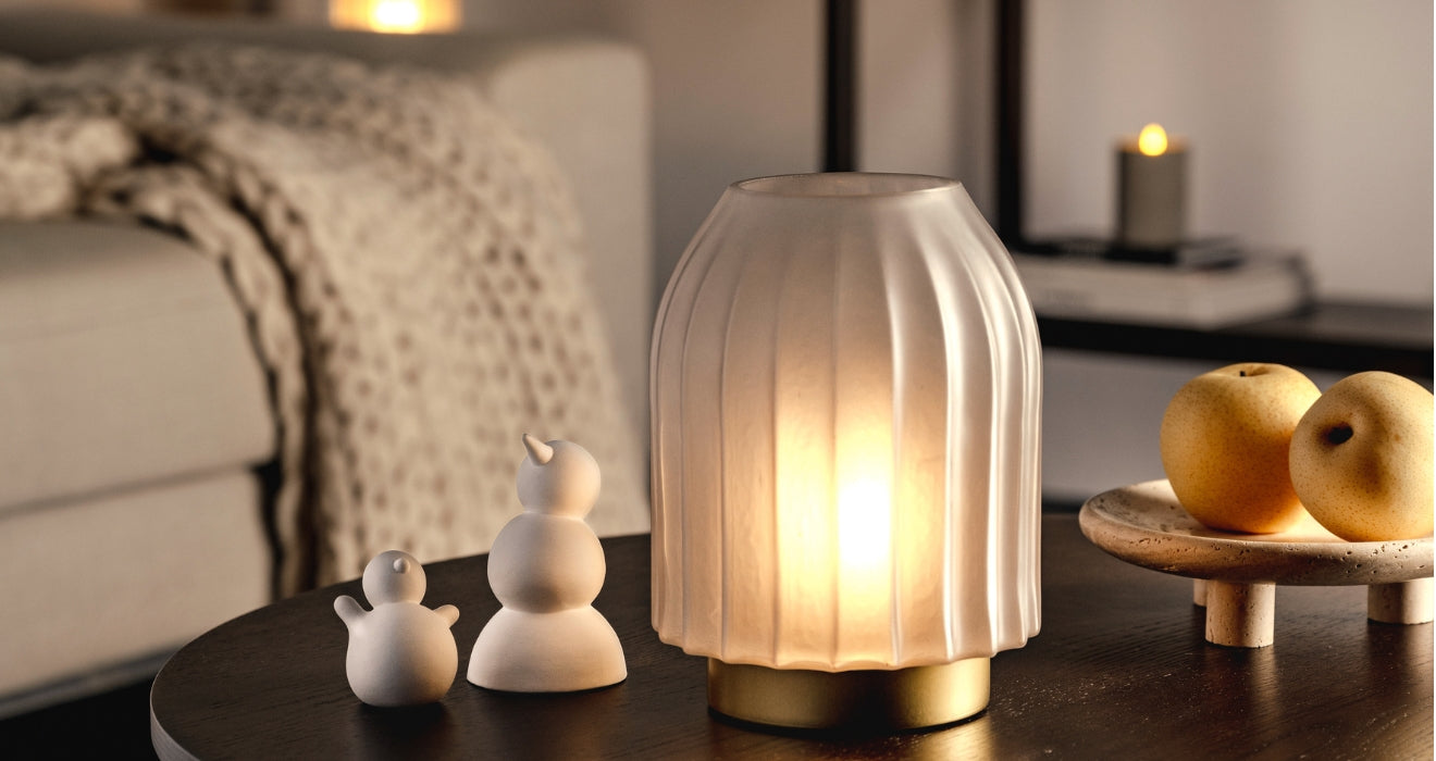 Eine stilvolle Glasleuchte mit geriffelter Oberfläche und goldenem Fuß sorgt für warmes Licht auf einem Tisch, dekoriert mit kleinen weißen Figuren und frischen Äpfeln.