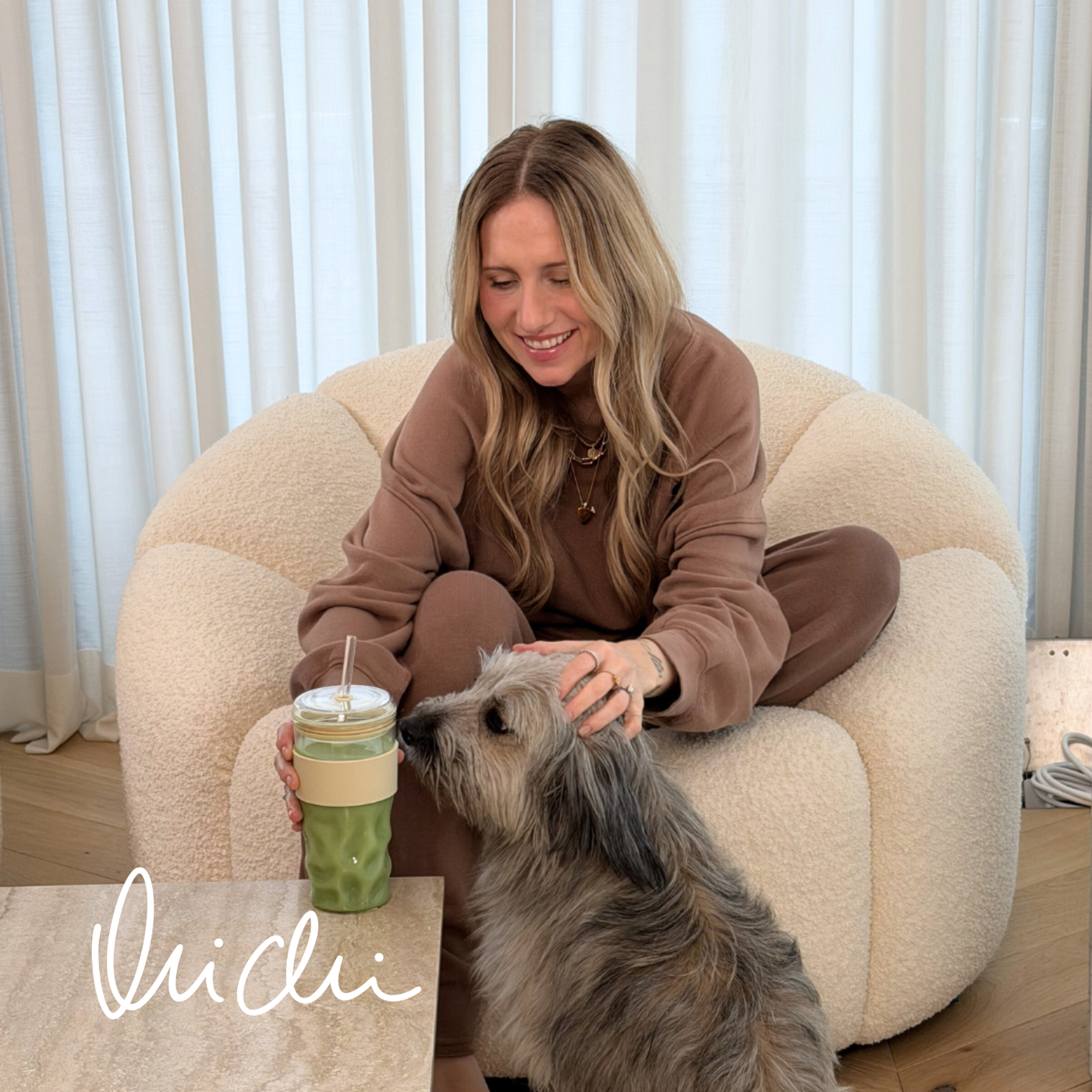 Frau im braunen Lounge-Set sitzt in einem cremefarbenen Sessel, streichelt einen kleinen Hund und hält einen Becher mit grünem Getränk; unten links steht „Michi“.