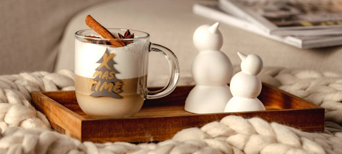 Eine gemütliche Szene auf einer Couch: Auf einem Holztablett steht eine Tasse Latte Macchiato mit Zimtstange und Sternanis, mit der Aufschrift „XMAS TIME“. Daneben zwei kleine weiße Schneemannfiguren aus Keramik, im Hintergrund ein aufgeschlagenes Magazin und eine dicke Strickdecke.