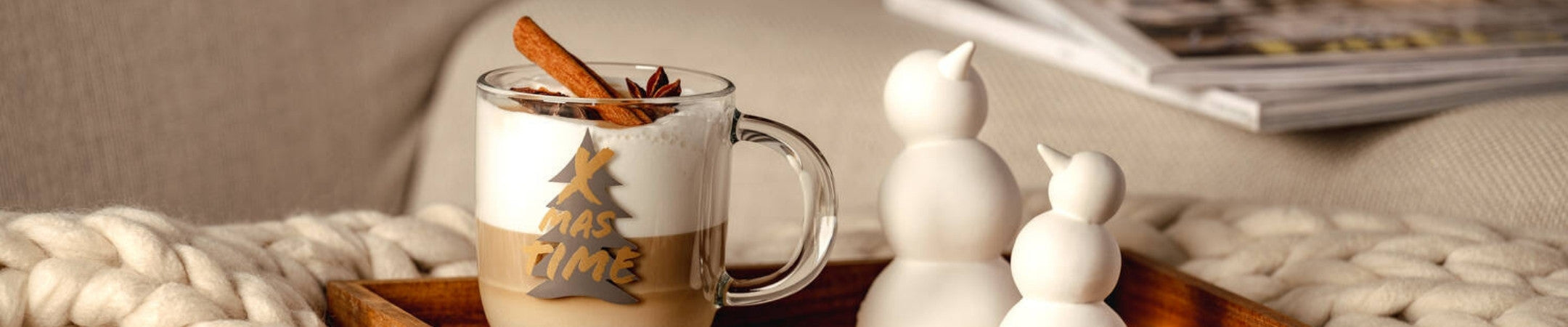 Eine weihnachtlich-gemütliche Nahaufnahme: Auf einem Holztablett steht ein Glas Latte Macchiato mit Zimtstange und Sternanis, daneben zwei weiße Schneemannfiguren. Im Hintergrund liegen Zeitschriften auf einem Sofa mit dicker Strickdecke.