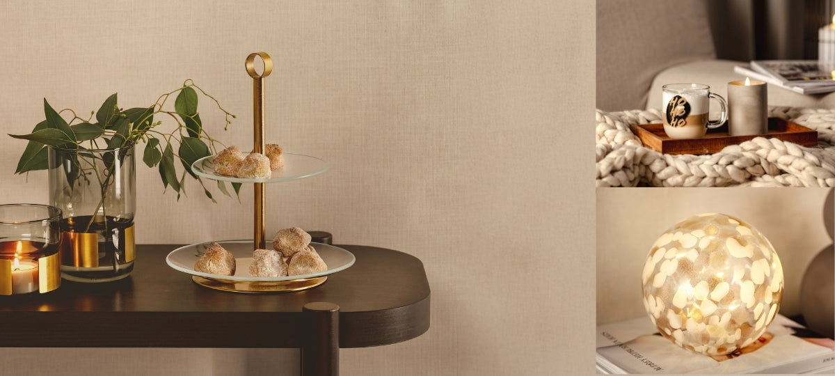 Eine goldene Etagere mit Gebäckstücken steht auf einem dunklen Tisch neben gläsernen Windlichtern und einer Vase mit grünen Zweigen, daneben sind dekorative Wohnaccessoires wie eine Kerze im Betonlook, eine leuchtende Glaskugel und ein Tablett mit Tasse arrangiert.