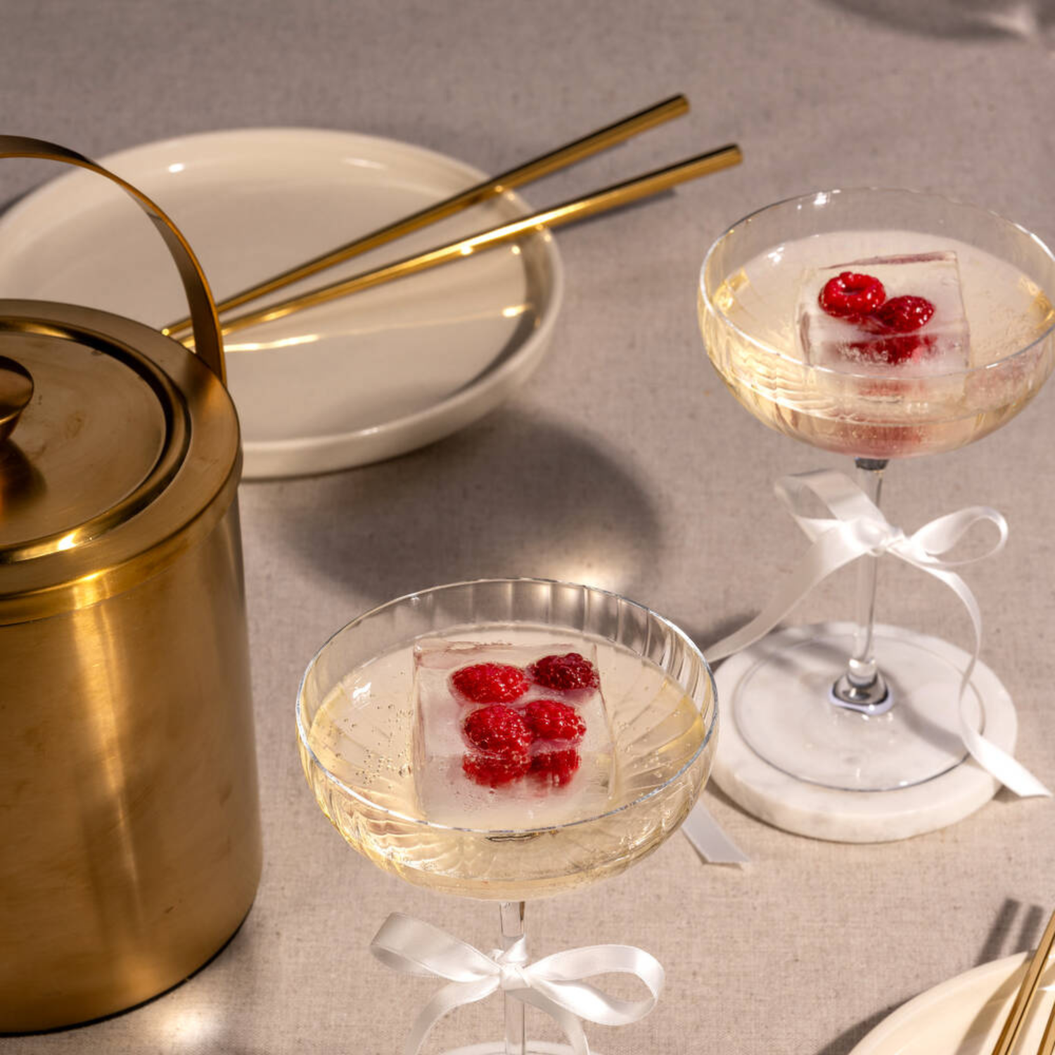 Zwei zarte Champagnerschalen mit gefrorenen Himbeeren auf großen Eiswürfeln sind mit weißen Schleifen dekoriert und begleiten ein stilvoll gedecktes Tischarrangement.