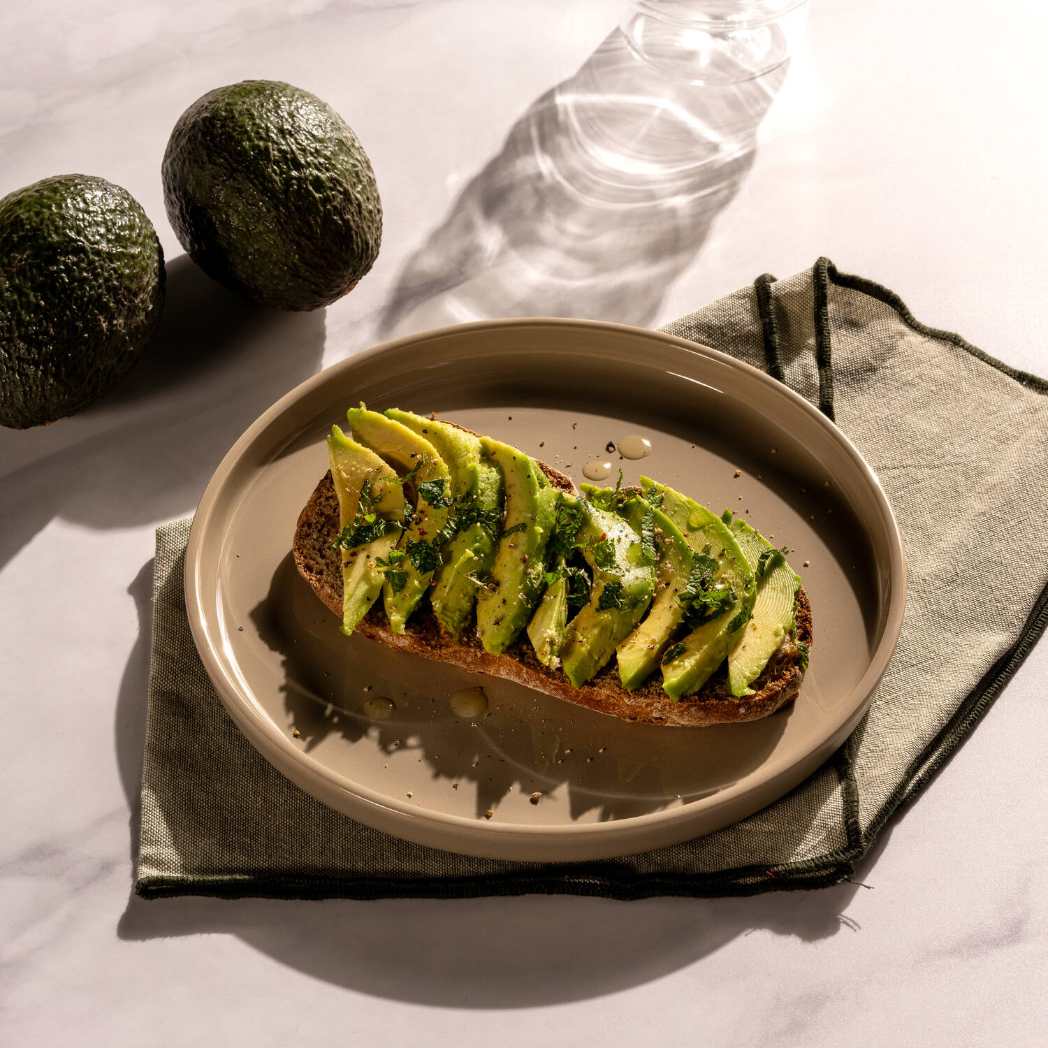 Ein kunstvoll belegtes Avocadobrot auf Vollkorn liegt auf einem hellen Teller mit Stoffserviette – daneben runden zwei Avocados das Bild stilvoll ab.