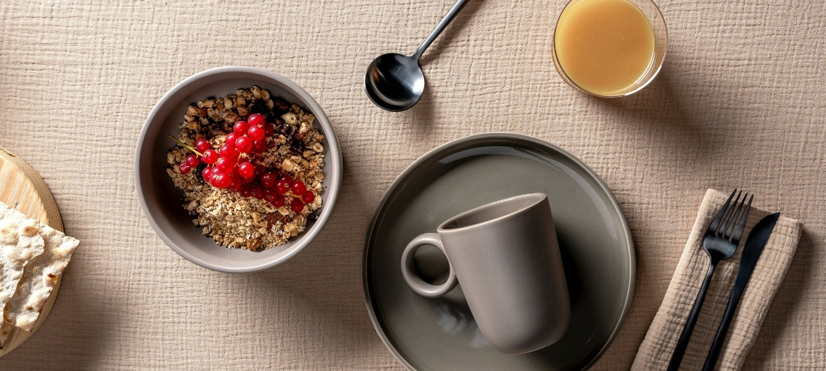 Frühstückstisch mit Müslischale, grauem Keramikbecher, Saft und Besteck auf beigem Untergrund