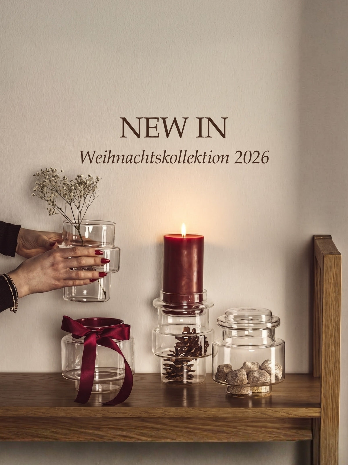 NEW IN Weihnachtskollektion 2026. Glasdosen mit Kerze, Trockenblumen und Dekoelementen auf Holzregal, warme festliche Atmosphäre