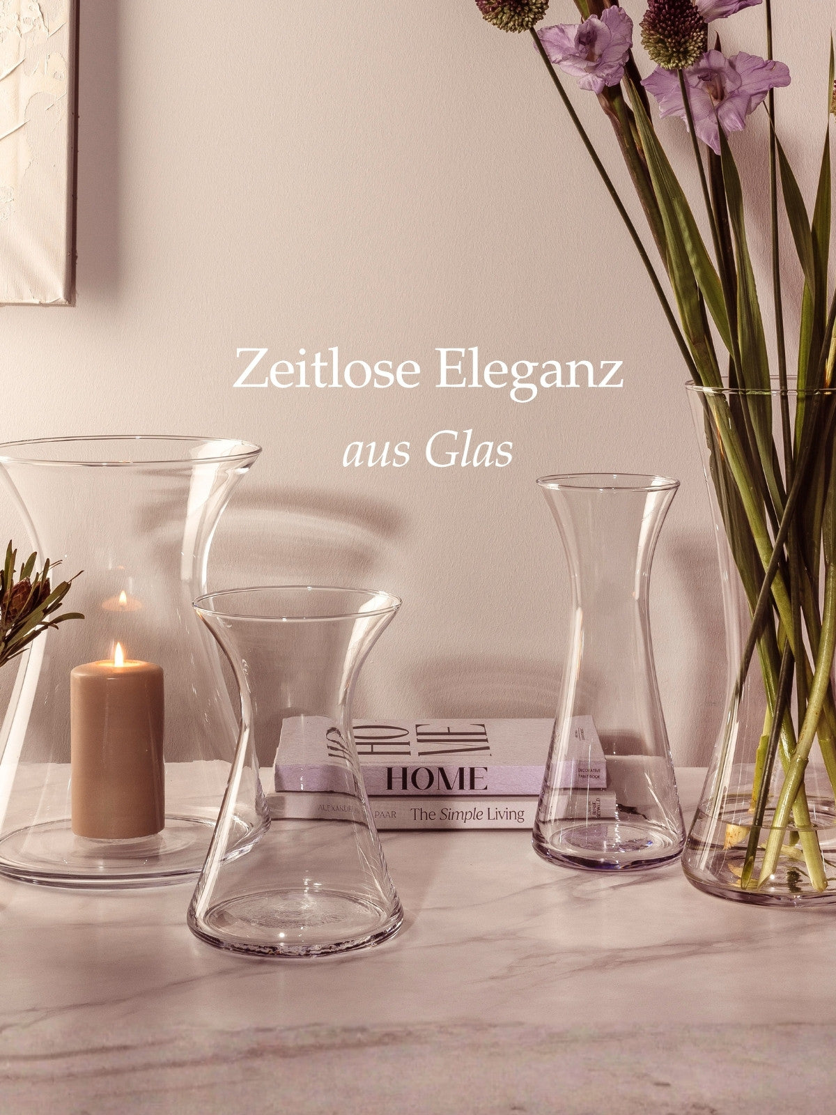 Glasvasen in unterschiedlichen Formen stehen dekorativ auf einer Marmorfläche, kombiniert mit einer Kerze im Windlicht, Büchern und hohen Blumen, begleitet vom Schriftzug „Zeitlose Eleganz aus Glas“.