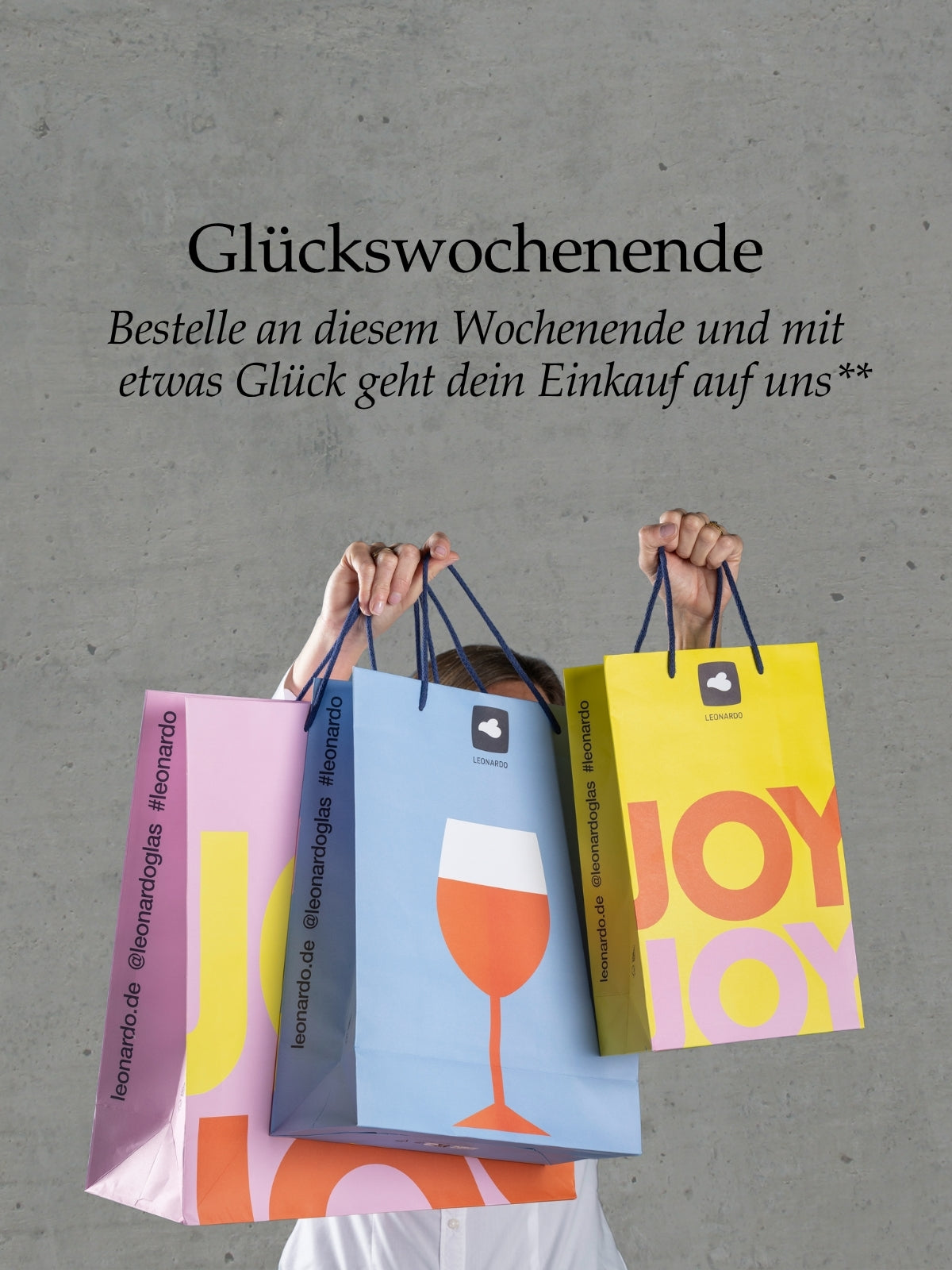 Glückswochenende: Bunte LEONARDO Papiertüten mit Weinglas- und Joy-Motiven in Händen, grauer Betonhintergrund