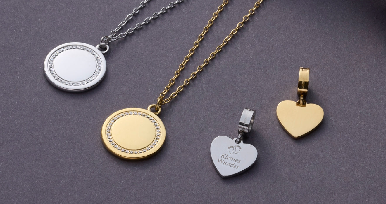 Zwei Ketten mit runden Anhängern und drei herzförmige Gravur-Plättchen in Silber und Gold liegen auf grauem Hintergrund – einer davon trägt die Gravur „Kleines Wunder“ mit Babyfüßchen.