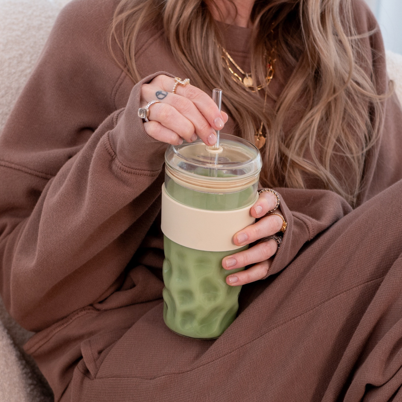 Wiederverwendbarer Glas-Cup mit grünem Getränk und Glasstrohhalm, gehalten von einer sitzenden Person in brauner Kleidung.