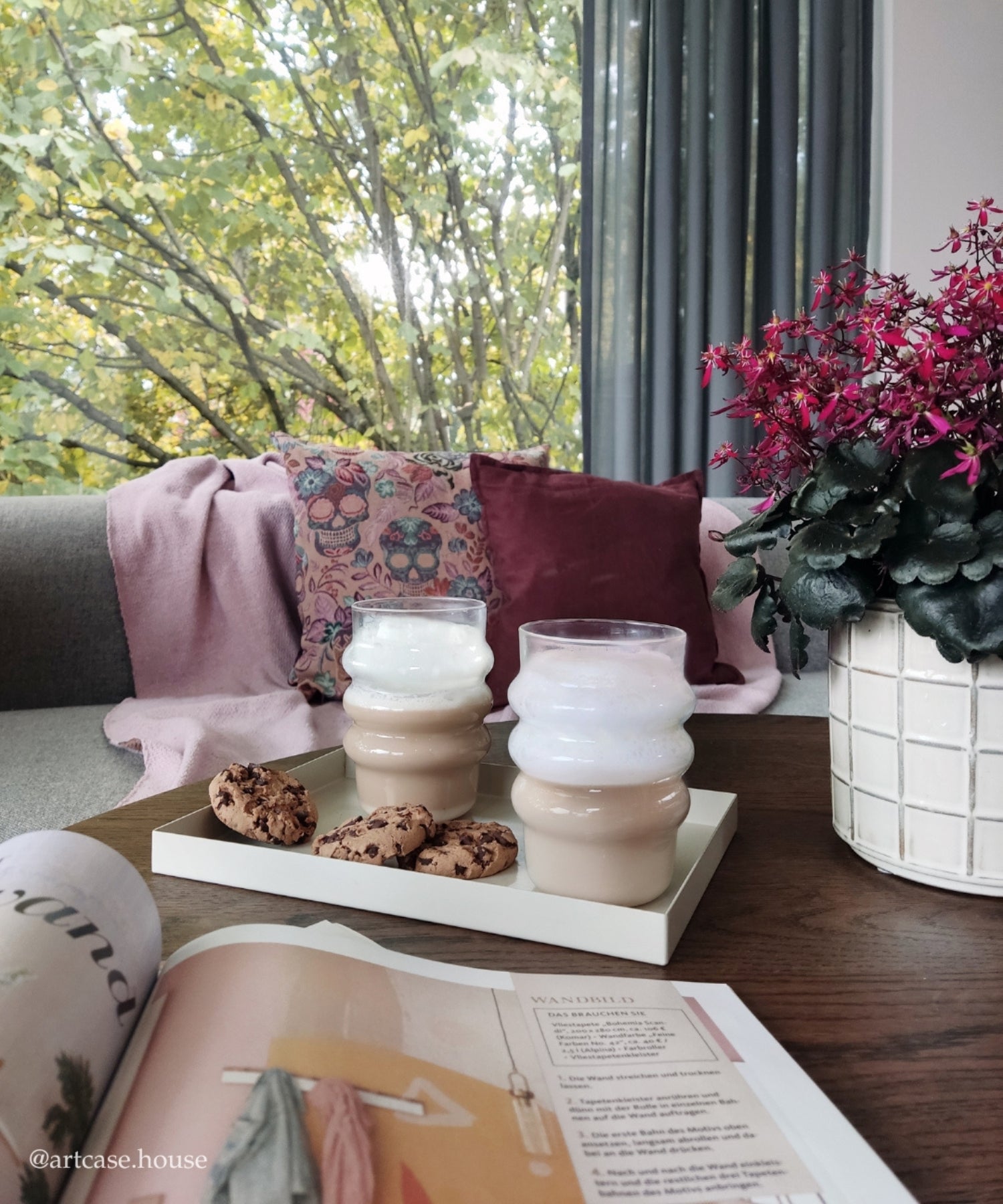 Zwei Milchkaffees in rundlich geformten Gläsern auf einem Tablett mit Schokokeksen, gemütliche Atmosphäre mit Sofakissen und Kuscheldecke am Fenster.