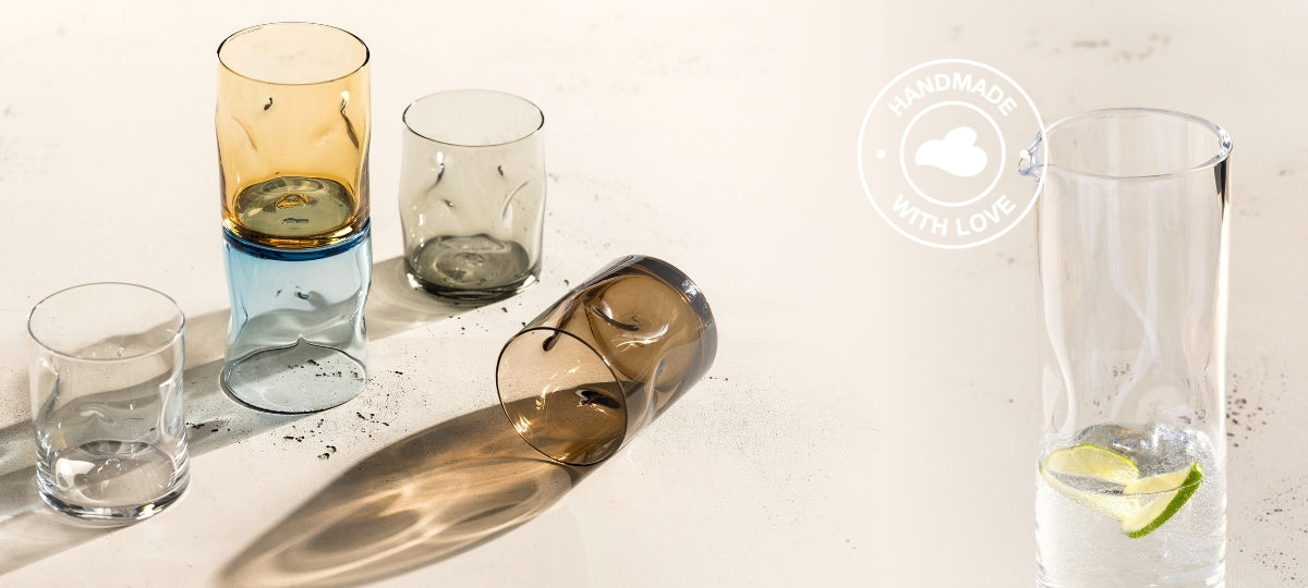 Fünf farbige Trinkgläser in Blau, Gelb, Braun, Grau und Transparent mit organischer Struktur, arrangiert neben einer schlichten Glaskaraffe mit Zitronen- und Limettenscheiben im Wasser, beleuchtet von warmem Sonnenlicht.