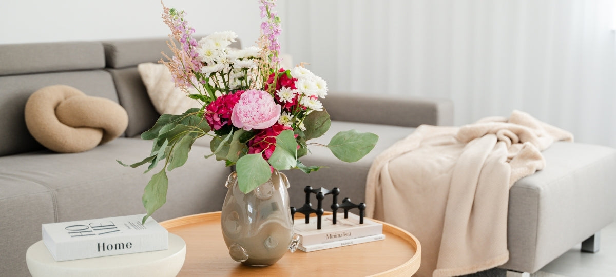 Elegante Glasvase mit buntem Blumenstrauß auf Holztisch neben Sofa in heller Wohnzimmeratmosphäre