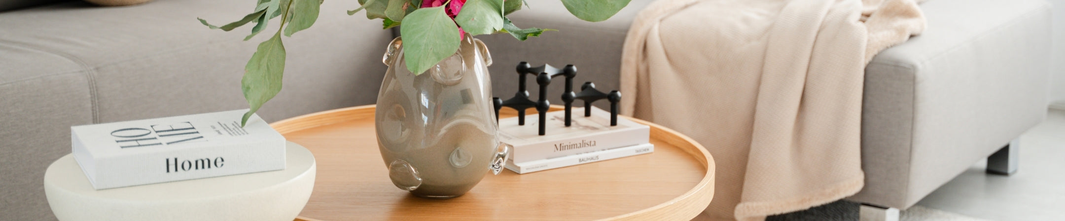 Elegante beige Vase neben Deko-Objekten auf rundem Holztisch, neben grauem Sofa mit Kissen und Decke