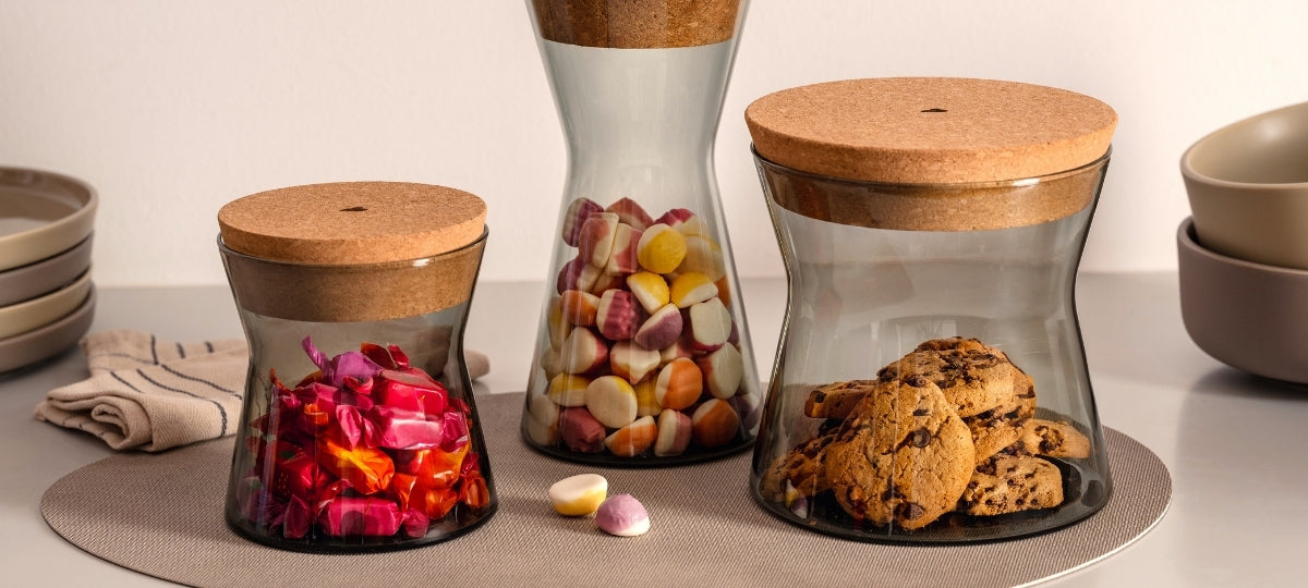 Verschiedene Vorratsdosen aus Glas mit Korkdeckeln gefüllt mit getrockneten Blütenblättern, Bonbons und Keksen auf beigem Untergrund