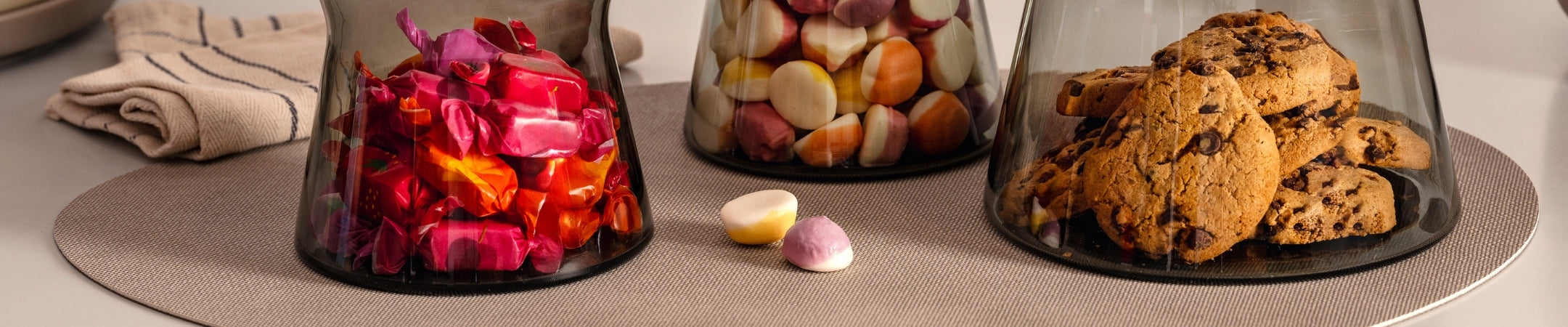 Verschiedene Vorratsdosen aus Glas mit Inhalt: rote Gummibärchen, Nüsse, Kekse mit Schokoladenstücken auf beigefarbenem Untergrund