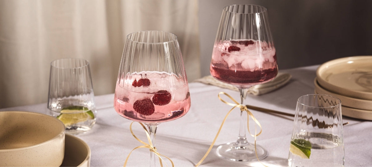 Elegante Weingläser mit geripptem Design, gefüllt mit rosa Getränk und roten Beeren, auf gedecktem Tisch mit Tellern und Besteck
