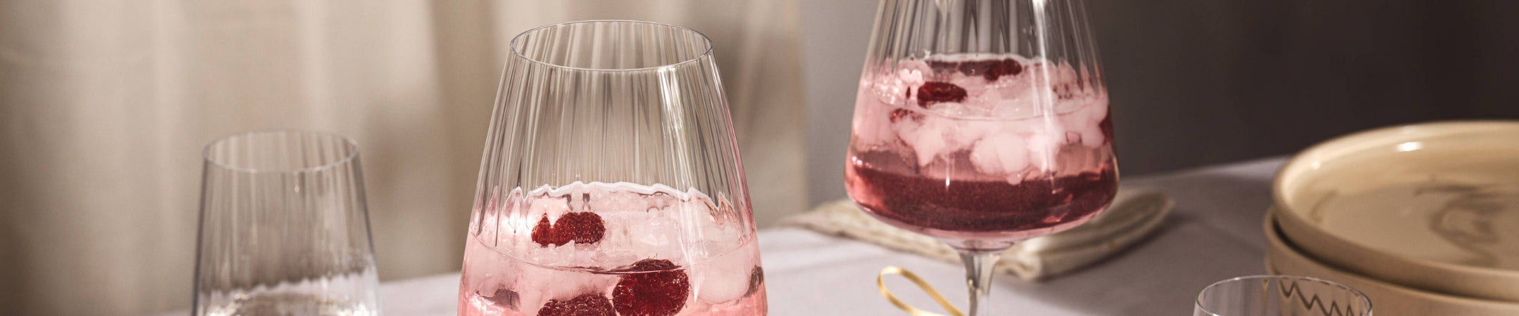 Drei Weingläser mit geriffelter Oberfläche, gefüllt mit rosafarbenem Wein und Eiswürfeln, auf hellem Tisch mit Tellern im Hintergrund