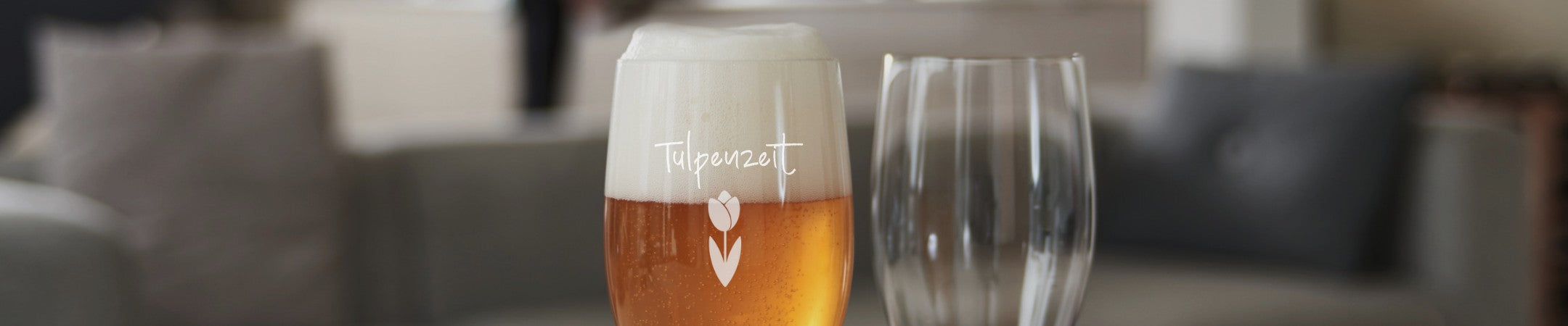 LEONARDO Weizenglas mit Gravur „Tulpenzeit“ und Tulpenmotiv, gefüllt mit frisch gezapftem Bier, daneben ein weiteres klares Glas – stilvoll inszeniert auf einer gemütlichen Wohnzimmercouch.
