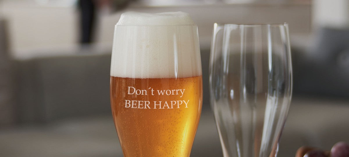 LEONARDO Weizenglas mit Gravur „Don’t worry BEER HAPPY“, neben einem weiteren schlichten Bierglas auf gemütlicher Wohnzimmerkulisse.