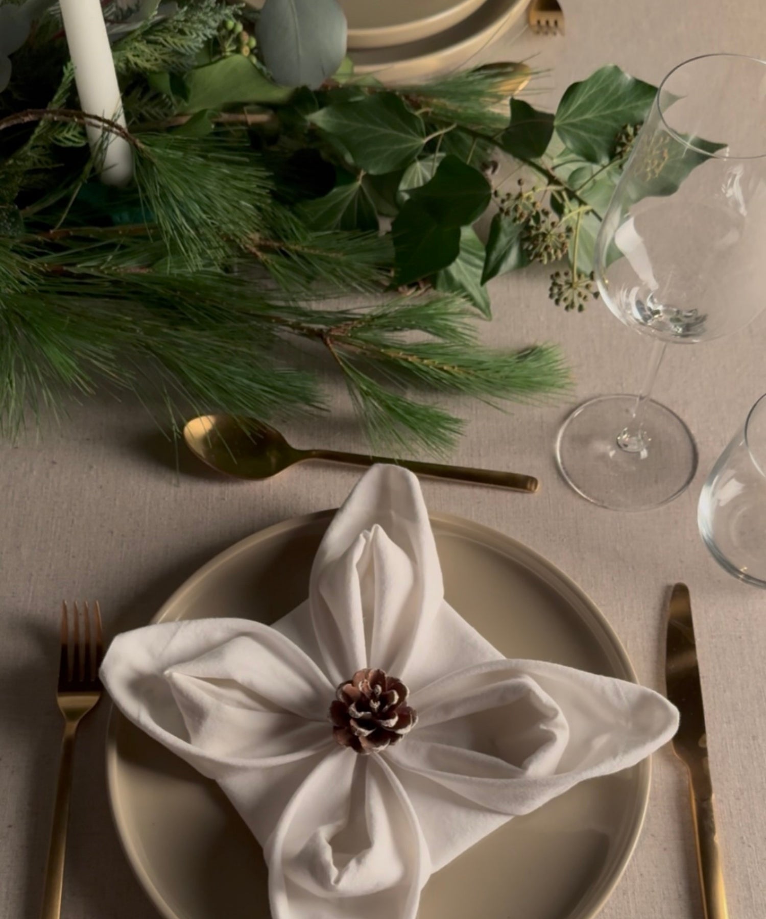 Weihnachtlich gedeckter Tisch mit beigem Geschirr, goldfarbenem Besteck, Weingläsern und einem weißen Stoffserviettenstern, der in der Mitte mit einem Tannenzapfen fixiert ist; darüber ein üppiger grüner Kranz aus Tannenzweigen und Blättern.