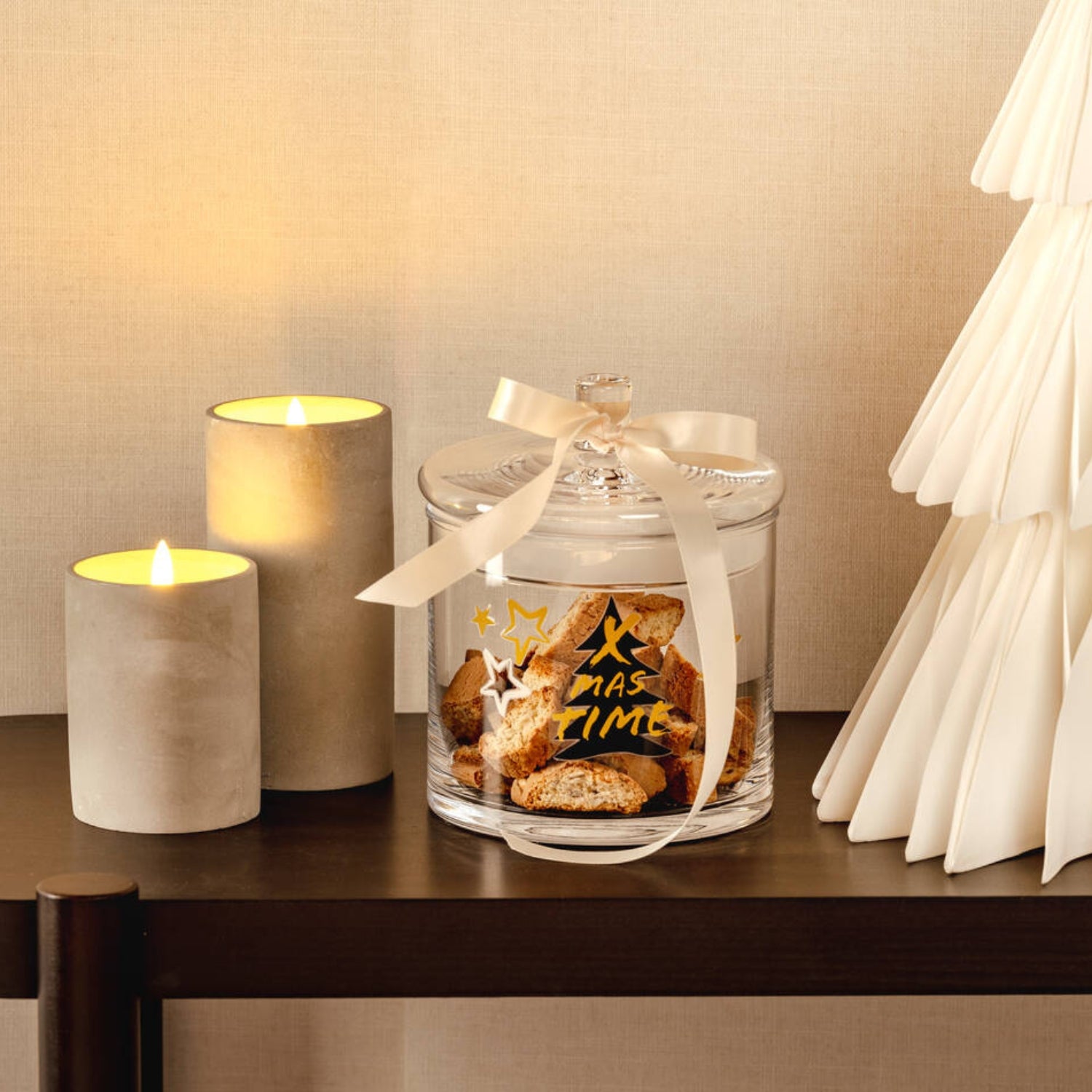 Ein dekoratives Glas mit goldfarbenem Weihnachtsmotiv ist mit Gebäck gefüllt und mit einer Schleife verziert, daneben stehen zwei LED-Kerzen.